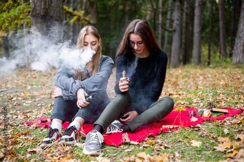 Vape lgbt teenagers. Bisexual lesbian young caucasian teenage girls in casual clothes vape electronic cigarette on the street in the park in the autumn evening. Bad habit.