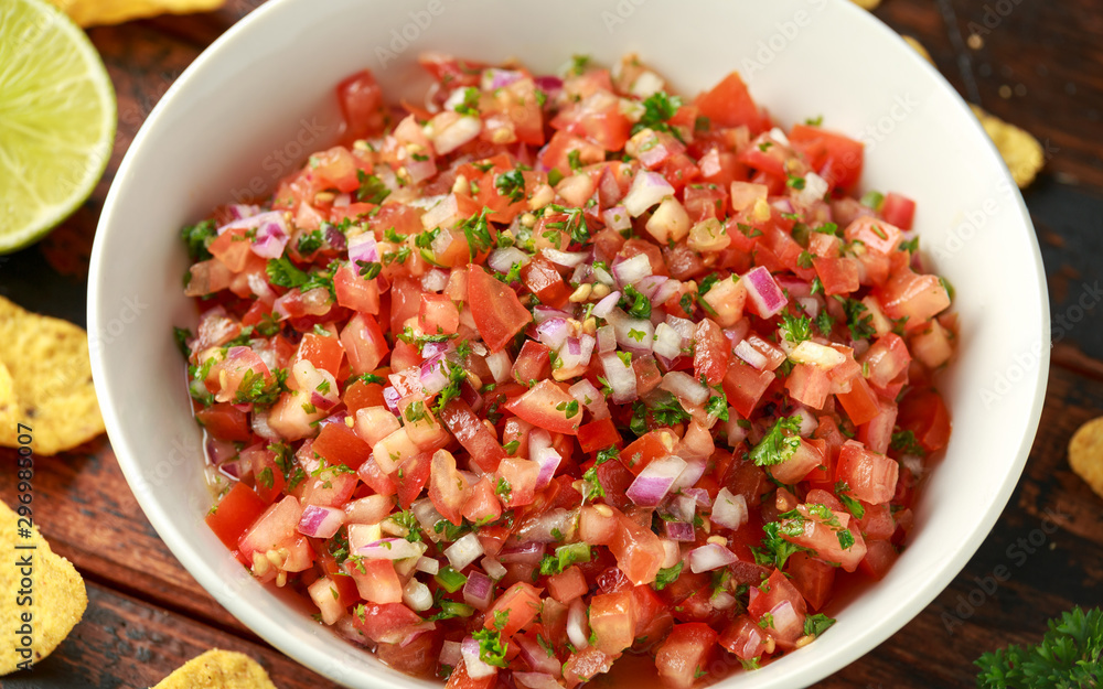 Mexican Tomato Salsa in white bowl with lime, red onion, jalapeno pepper, parsley and tortilla chips on wooden table Mexican Tomato Salsa in white bowl with lime, red onion, jalapeno pepper, parsley and tortilla chips on wooden table