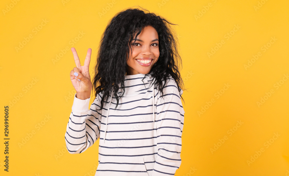 Striped mood. Playful African American girl in a striped hoodie is posing with a broad smile on the yellow background, looking to the camera and showing a V-sign with her right hand. Striped mood. Playful African American girl in a striped hoodie is posing with a broad smile on the yellow background, looking to the camera and showing a V-sign with her right hand.