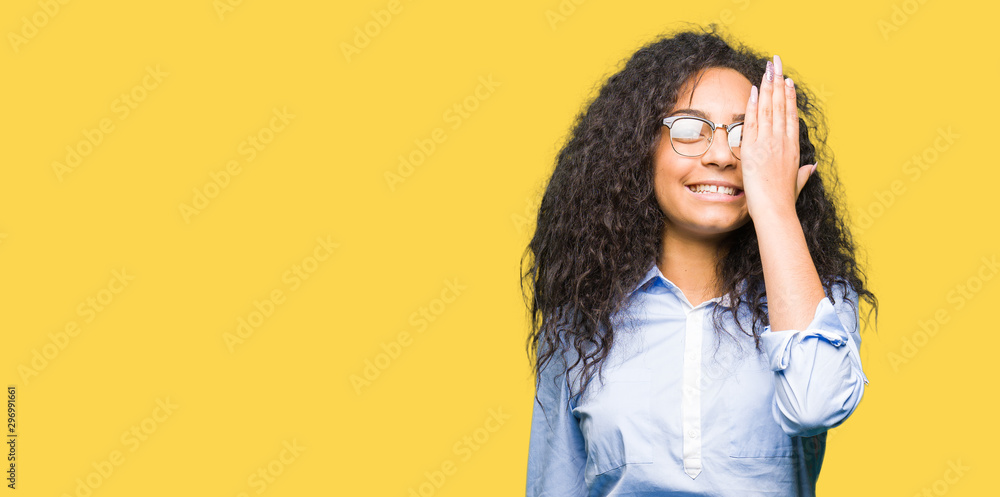 Young beautiful business girl with curly hair wearing glasses covering one eye with hand with confident smile on face and surprise emotion. Young beautiful business girl with curly hair wearing glasses covering one eye with hand with confident smile on face and surprise emotion.
