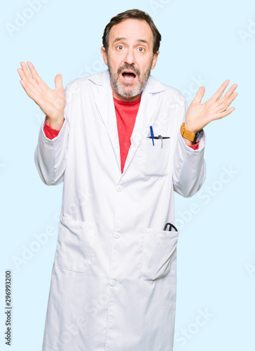 Middle age therapist wearing white coat clueless and confused expression with arms and hands raised. Doubt concept.