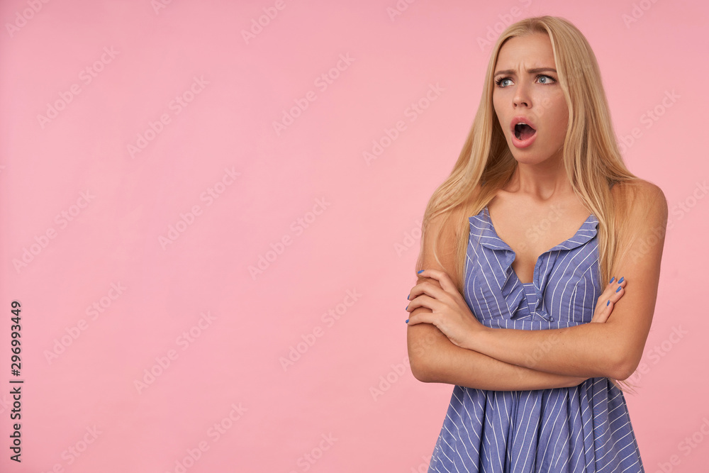 Shocked young blonde lady with casual hairstyle wearing blue summer dress over pink background, looking aside with wide mouth opened and keeping hands folded Shocked young blonde lady with casual hairstyle wearing blue summer dress over pink background, looking aside with wide mouth opened and keeping hands folded