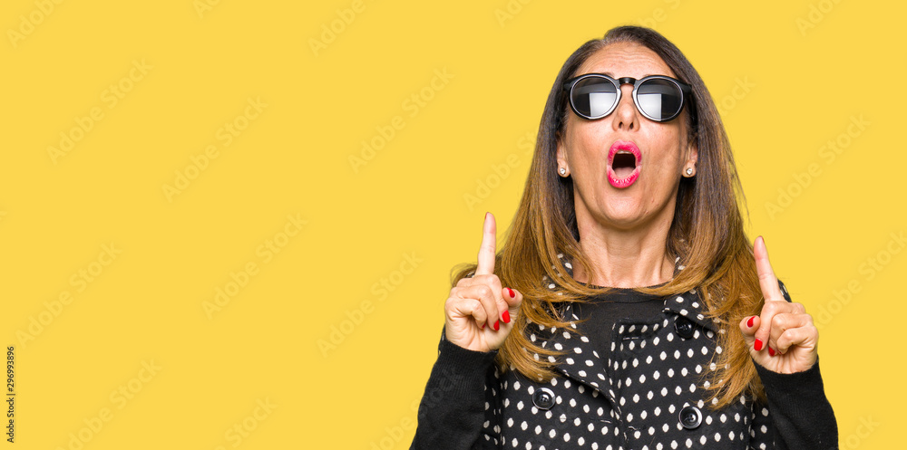 Obraz premium Beautiful middle age woman wearing sunglasses amazed and surprised looking up and pointing with fingers and raised arms.