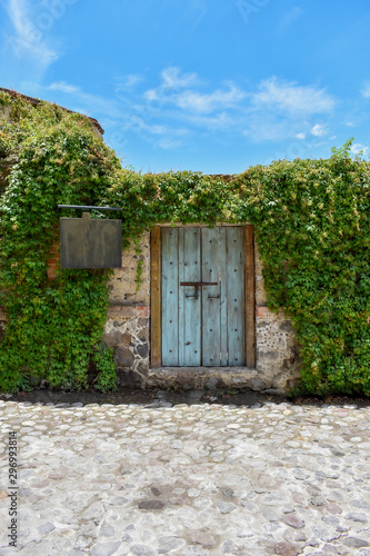 Fachada de casa de pueblo con puerta rústica de madera pintada de color azul con cerradura antigua, con un letrero de madera, rodeada del enredadera verde y con cielo azul al fondo photo