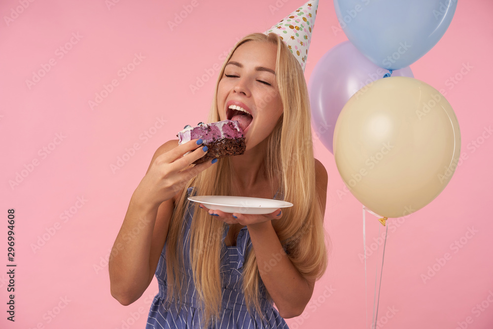 Portrait of young delighted blonde female in holiday outfit holding big piece of delicious cake and going to eat it, celebrating birthday with multicolored air balloons, posing over pink background Portrait of young delighted blonde female in holiday outfit holding big piece of delicious cake and going to eat it, celebrating birthday with multicolored air balloons, posing over pink background