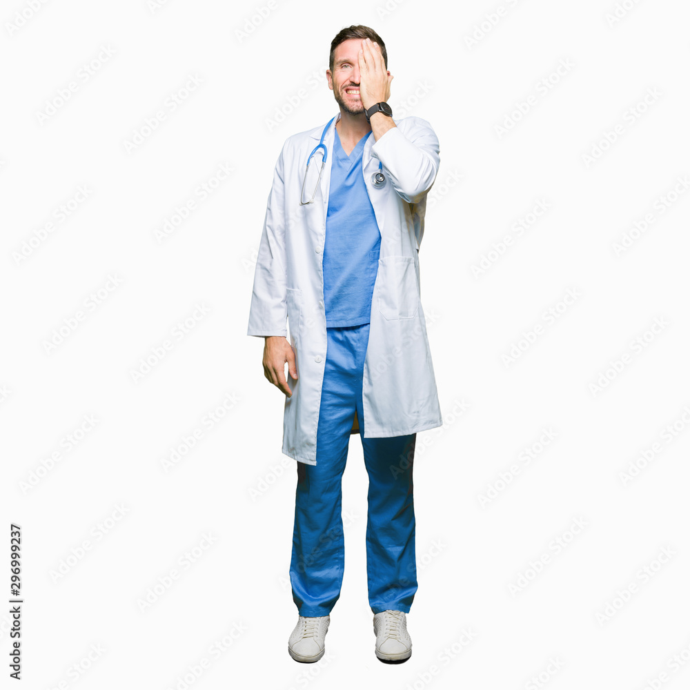 Handsome doctor man wearing medical uniform over isolated background covering one eye with hand with confident smile on face and surprise emotion. Handsome doctor man wearing medical uniform over isolated background covering one eye with hand with confident smile on face and surprise emotion.