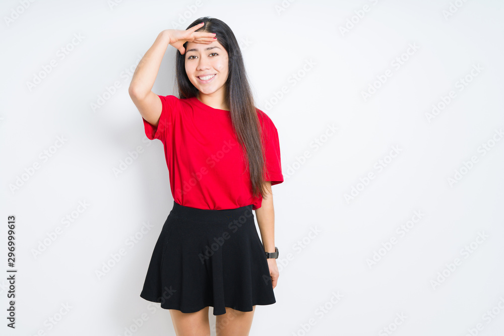 Beautiful brunette woman wearing red t-shirt and skirt over isolated background very happy and smiling looking far away with hand over head. Searching concept. Beautiful brunette woman wearing red t-shirt and skirt over isolated background very happy and smiling looking far away with hand over head. Searching concept.