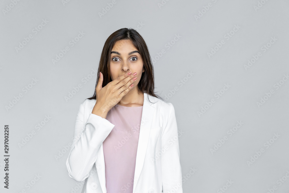 Excited surpised young adult indian business lady in formal wear white suit looking at camera cover mouth with hand. Shocked businesswoman shopper standing isolated on grey background with copy space Excited surpised young adult indian business lady in formal wear white suit looking at camera cover mouth with hand. Shocked businesswoman shopper standing isolated on grey background with copy space