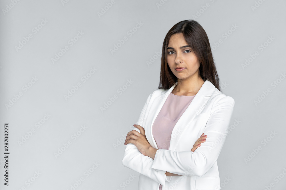 Serious young adult indian businesswoman in white formal wear crossed arm on chest and looking at camera. Woman standing isolated on grey background with copy space Serious young adult indian businesswoman in white formal wear crossed arm on chest and looking at camera. Woman standing isolated on grey background with copy space