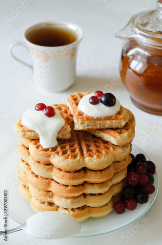 A pile of freshly baked homemade waffles sprinkled with crean and with berries on the top on the table and tea utensils in the background