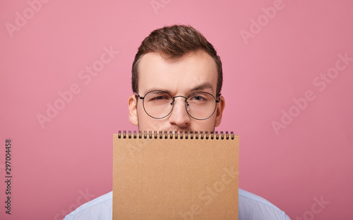 Fotografija A young student in a gently blue shirt with glasses covered his face with a brown loose-leaf notebook and looks into the frame, is on a pink background