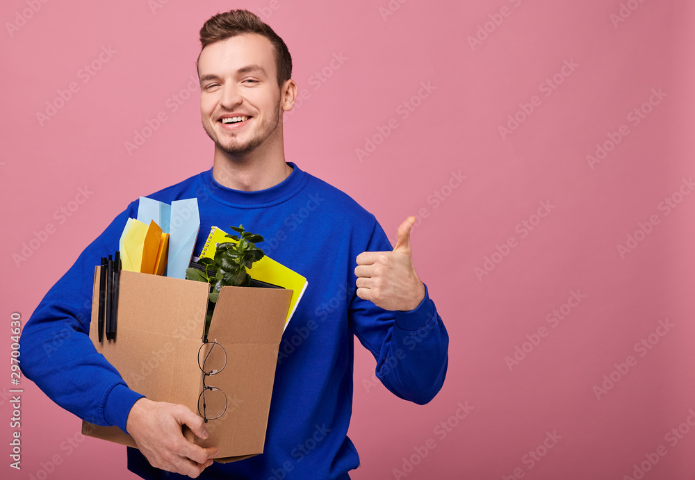 Focus on the box. A cool stylish retro guy in a dark blue sweater is standing hugging a box with green plant, yelow notebook and paper airplane with hands. The guy was lose work. Focus on the box. A cool stylish retro guy in a dark blue sweater is standing hugging a box with green plant, yelow notebook and paper airplane with hands. The guy was lose work.