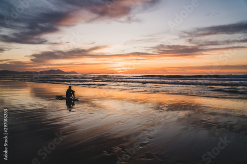 Silhouette of a surfer sitting on the beach of the Atlantic Ocean, near San Sebastian and Bilbao, North of Spain, watching a dramatic stunning colorful sunset