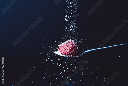 Sprinkling of sugar into spoon with fresh strawberry against dark background