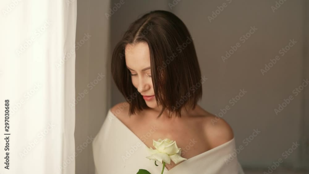 Closeup portrait of beautiful woman with white rose in white interior. Young attractive confident brunette lady at the window. Female natural beauty. Slow motion video Closeup portrait of beautiful woman with white rose in white interior. Young attractive confident brunette lady at the window. Female natural beauty. Slow motion video