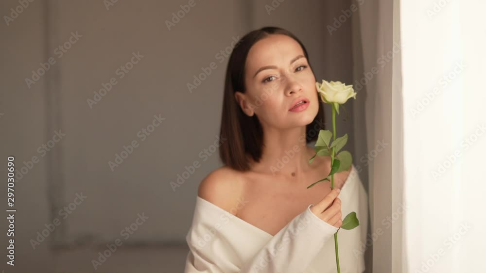 Closeup portrait of beautiful woman with white rose in white interior. Young attractive confident brunette lady at the window. Female natural beauty. Slow motion video Closeup portrait of beautiful woman with white rose in white interior. Young attractive confident brunette lady at the window. Female natural beauty. Slow motion video