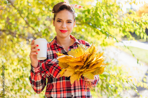 Beautiful woman with make up and hair in pin up style holding big bouquet of maple yellow leaves and white cup of coffee to go. photo