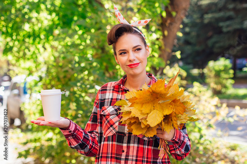 Beautiful woman with make up and hair in pin up style holding big bouquet of maple yellow leaves and white cup of coffee to go. photo