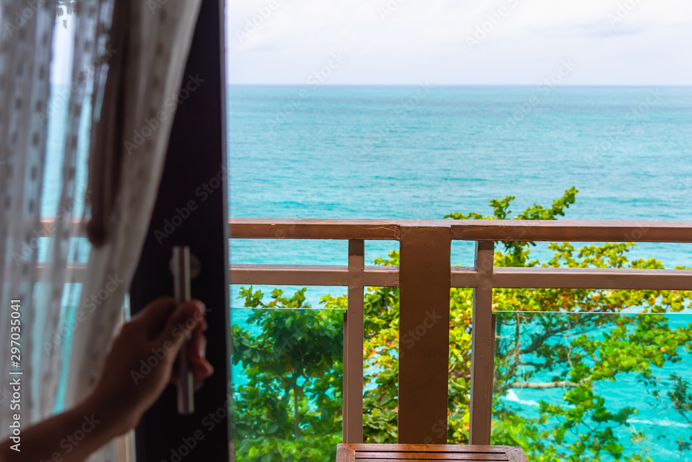 Woman's hand opening a glass door of hotel room to the ocean view balcony.