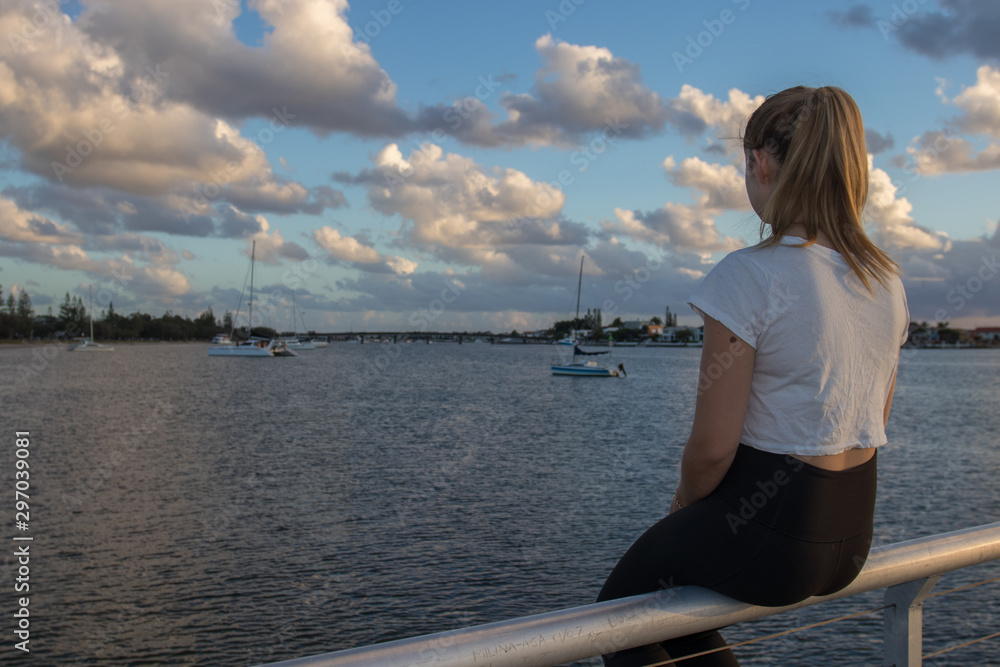 Blonde Woman Sitting On A Jetty Facing Towards Moored Boats On The Water At Sunrise