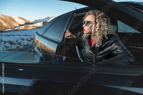 Fashion style wide angle young stylish attractive girl with bright red lips in a leather jacket and sunglasses sits in a retro 90s car with an open door on nature in the mountains at sunset © yanik88