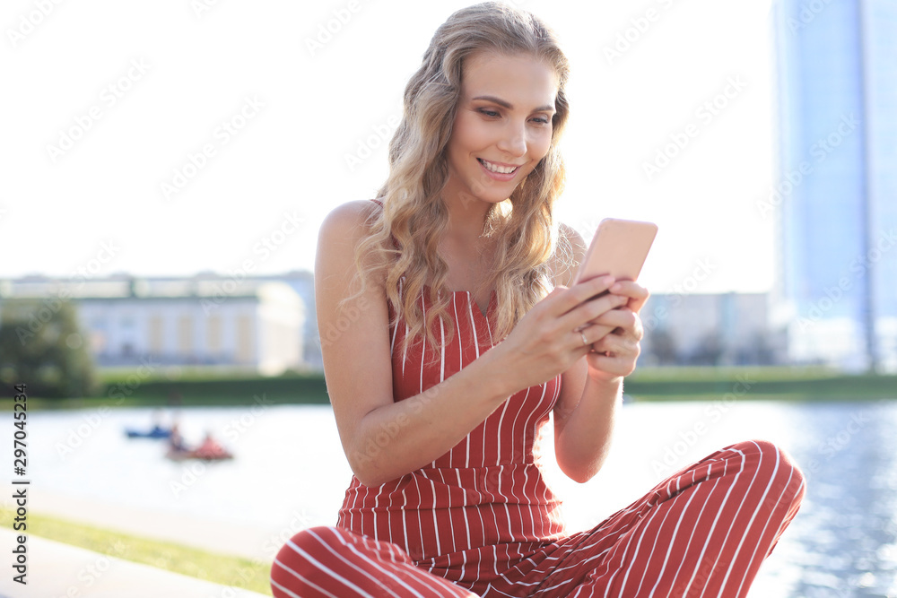 Portrait of pretty young woman sitting on riverbank with legs crossed during summer day, using smartphone