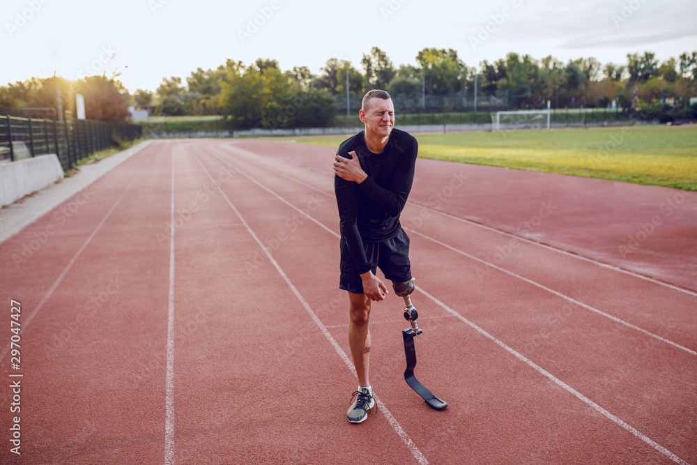 Sporty caucasian man with artificial leg standing on running track and holding painful arm. Sporty caucasian man with artificial leg standing on running track and holding painful arm.