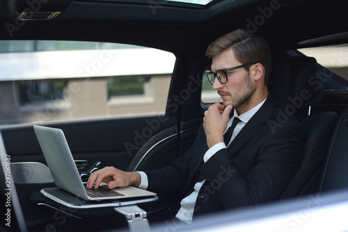 Thoughtful young businessman keeping hand on chin while sitting in the lux car and using his laptop. © ty