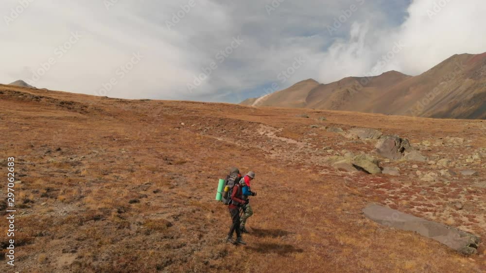 Aerial view of a couple of travelers man and woman with large backpacks in hats and sunglasses walk along an alpine plateau surrounded by epic mountains Aerial view of a couple of travelers man and woman with large backpacks in hats and sunglasses walk along an alpine plateau surrounded by epic mountains