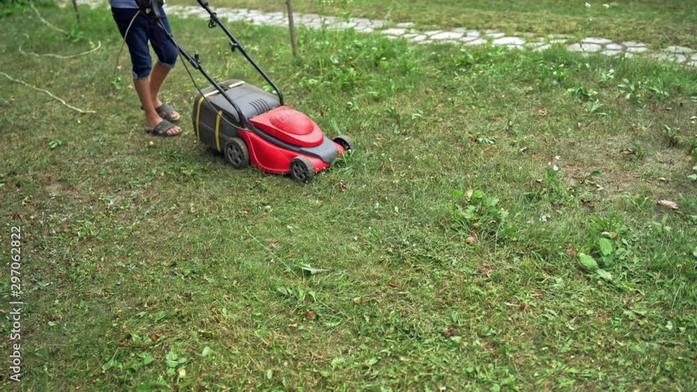 Electric lawn mower in the garden. Boy cutting grass by lawn mowing machine in summer day. Gardening activity. Electric lawn mower in the garden. Boy cutting grass by lawn mowing machine in summer day. Gardening activity.