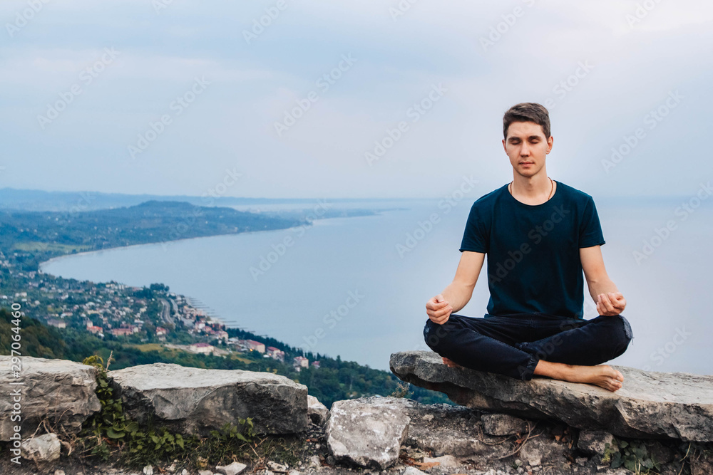Young handsome man in a meditative position sits against the background of the sea, mountains and city. Concept of freedom relaxation. Place for text or advertising