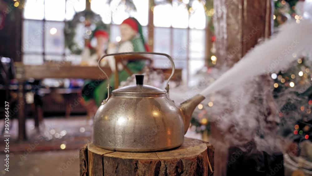 Boiling kettle in the room on blurred Christmas background. Hot kettle with boiled water and white steam on wooden log. Boiling kettle in the room on blurred Christmas background. Hot kettle with boiled water and white steam on wooden log.
