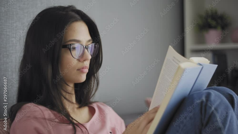Pretty beautiful happy female student with glasses reading a book on the couch of a modern house. Pretty beautiful happy female student with glasses reading a book on the couch of a modern house.