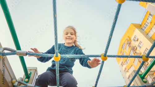 A charming little girl in casual clothes sits on the grid and looks at camera on playground in the urban district.