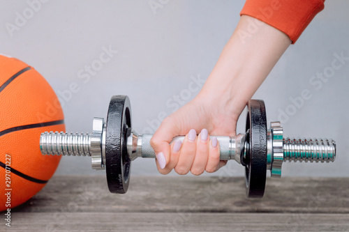 Woman's hand hold dumbbell near a basketball ball on a wooden bench. Fitness and outdoor workout.