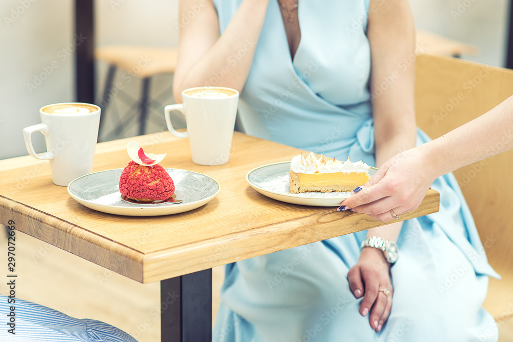 The female hand puts the piece of cake on the table at cafe. Woman hand is putting slice of cake on the table on background of female in blue dress. The female hand puts the piece of cake on the table at cafe. Woman hand is putting slice of cake on the table on background of female in blue dress.
