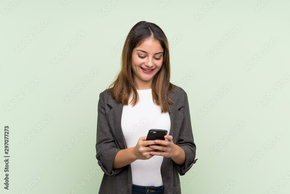 Young brunette girl with blazer over isolated green background sending a message with the mobile