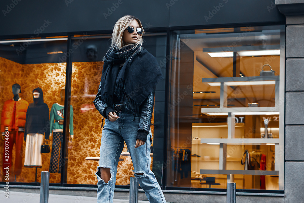 Fashion stylish woman in leather jacket, jeans, sweater and sunglasses walking on road on shops background. Elegant trendy outdoors portrait of pretty girl model on city street Fashion stylish woman in leather jacket, jeans, sweater and sunglasses walking on road on shops background. Elegant trendy outdoors portrait of pretty girl model on city street