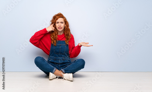 Redhead woman with overalls sitting on the floor making the gesture of madness putting finger on the head