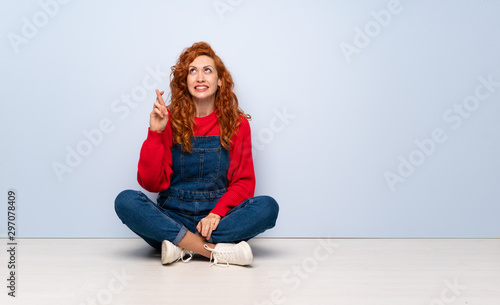 Redhead woman with overalls sitting on the floor with fingers crossing and wishing the best