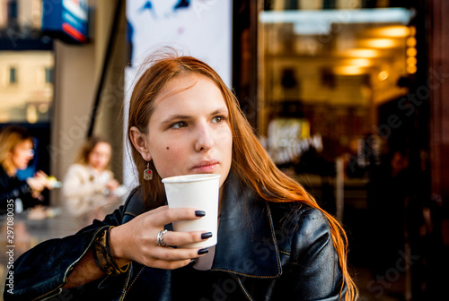 portrait of young teenager redhead girl with long hair with cup of hot coffee at city street cafe terrace