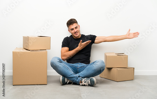 Handsome young man moving in new home among boxes extending hands to the side for inviting to come