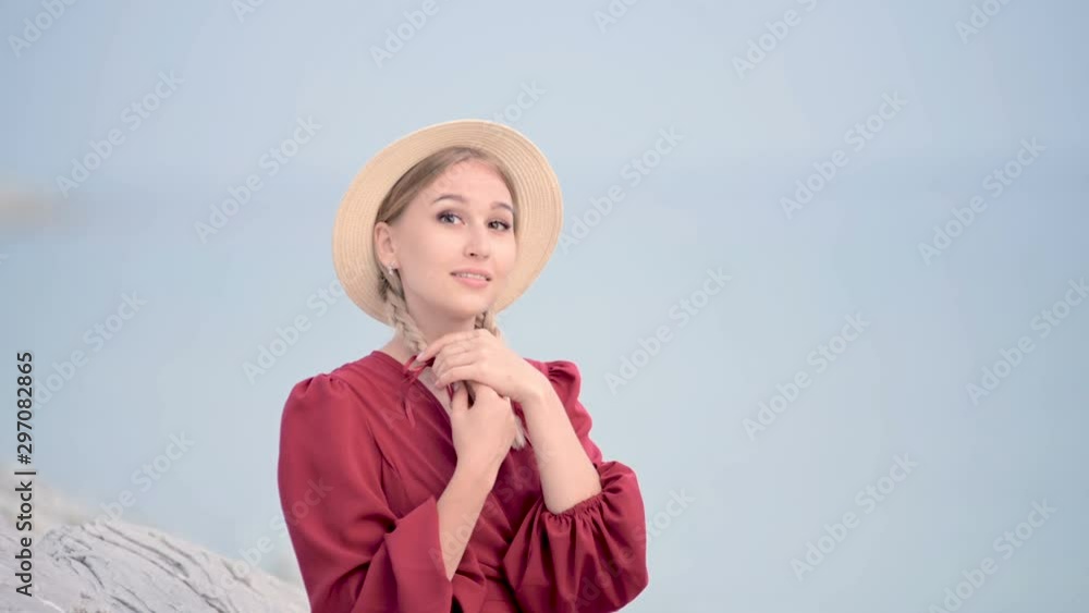 Portrait attractive caucasian young woman in a red dress and a straw hat with pigtails turns around sitting on the seashore smiling looking at the camera against the sea in defocus Portrait attractive caucasian young woman in a red dress and a straw hat with pigtails turns around sitting on the seashore smiling looking at the camera against the sea in defocus