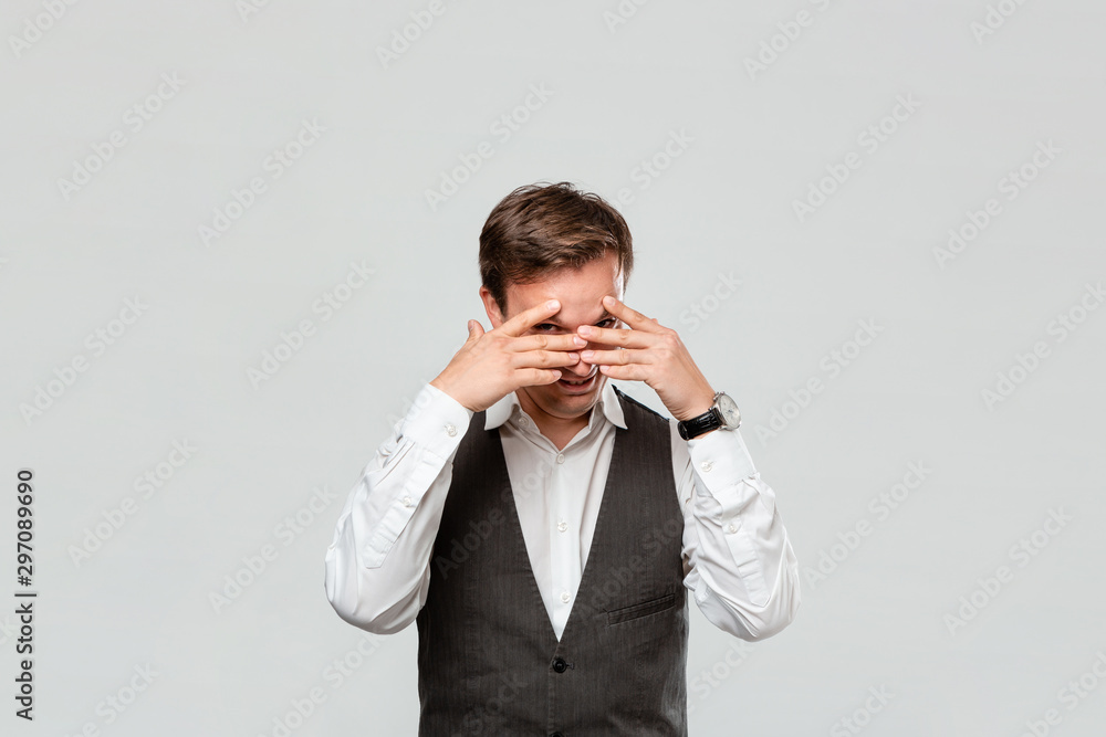 Handsome man in a white shirt and grey vest hiding behind his hands isolated over grey background. Handsome man in a white shirt and grey vest hiding behind his hands isolated over grey background.