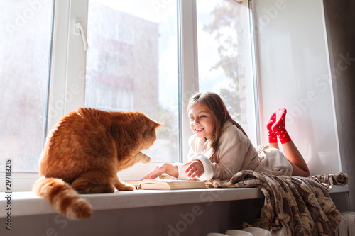 Quadro su tela Little girl in warm terry robe lying on window sill and reading a book