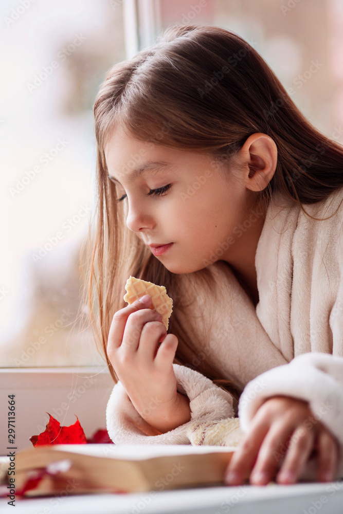 Cute little girl lying on the window, eating cookie and reading a book at home Cute little girl lying on the window, eating cookie and reading a book at home