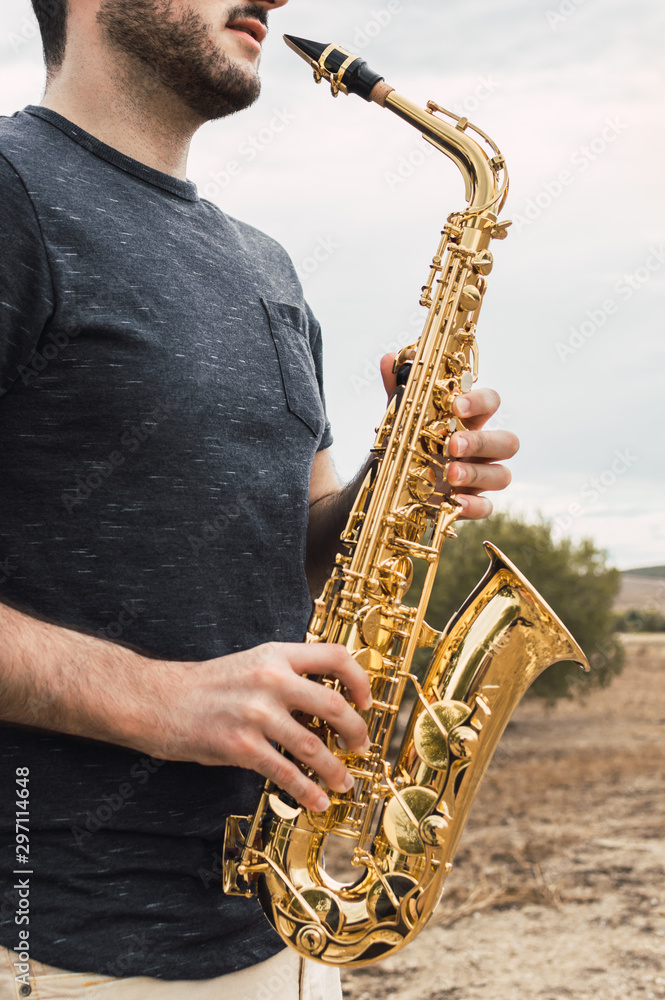 Fototapeta premium Young bearded man plays saxophone in a natural place during a cloudy day in the countryside. Playing musical instrument in the nature.