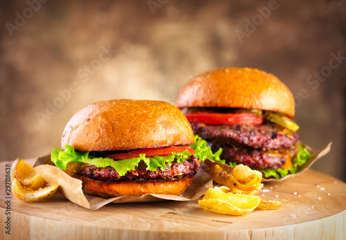 Hamburger and Double Cheeseburger with fries rotated on wooden table background. Cheeseburgers on fresh buns with succulent beef and fresh salad ingredients served with French Fries