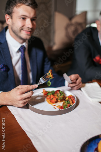 Impressive handsome young man eating baked lamb prepared with spicy sauce  keeps fork with small piece of meat  looks aside with pleasant smile. close up  restaurant concept