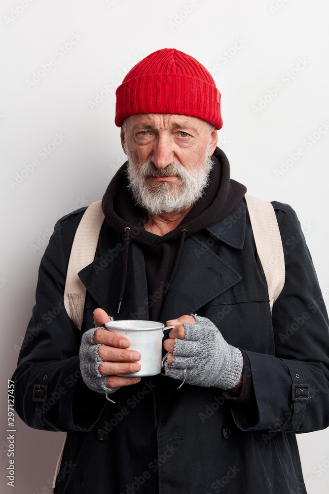 Homeless man wearing red hat and street clothes, look at camera with sadness. Beggar holding cup tea trying to warm up